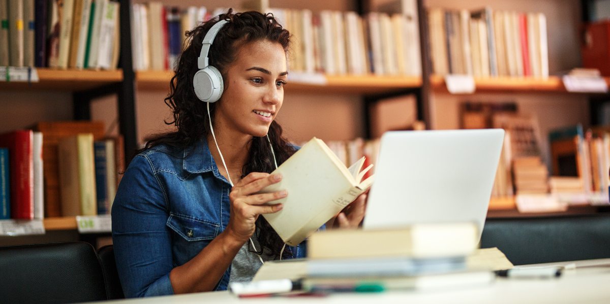 invatare online, femeie care invata in biblioteca si asista la curs online